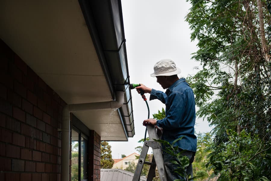 Person Standing on A Ladder Washing Gutters Using A Garden Hose