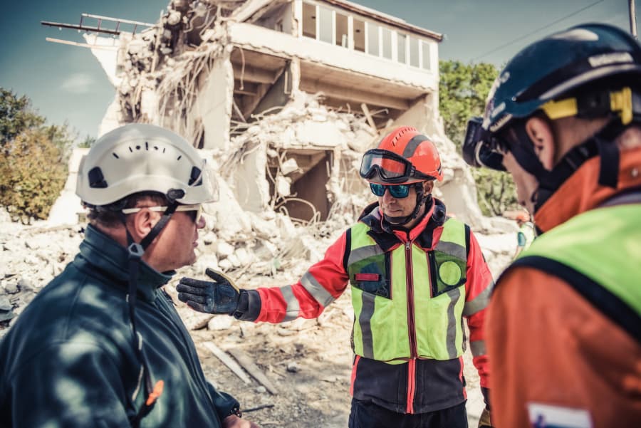 Three workers in hard hats discuss hurricane damage to destroyed building behind them