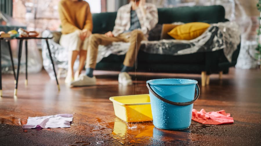 Water drips into buckets in living room while angry couple in background calls insurance company